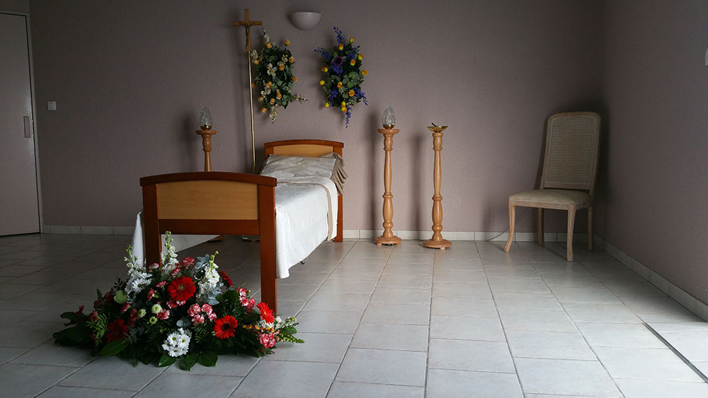 Chambre funéraire paisible avec lit vide pour défunt, arrangement floral au sol, croix, bougies et chaise. Atmosphère de recueillement et hommage.