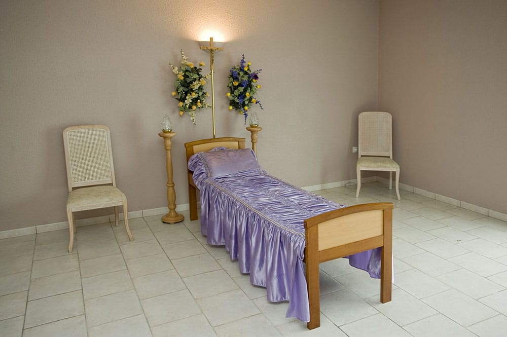 Chambre funéraire avec lit drapé de violet, deux chaises, décorations florales jaunes et bleues, et croix lumineuse.