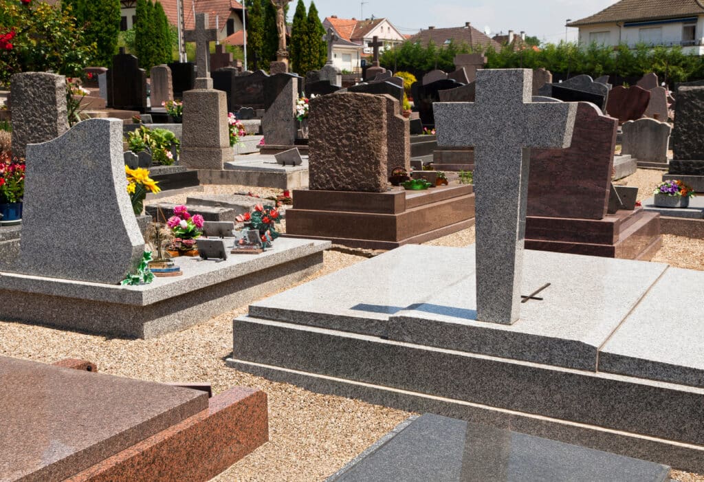 Cimetière ensoleillé avec de nombreuses tombes en granit de différentes formes et couleurs. Des croix et des bouquets de fleurs ornent les sépultures.