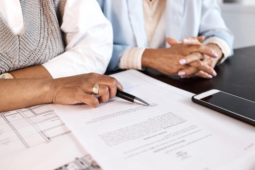 Deux femmes lors d'une réunion d'affaires, l'une tenant un stylo sur un contrat (mentionnant "1.2 Powers."), l'autre attentive. Plans architecturaux ("KITCHEN", "LIFT", "Existing Floor Slab") et téléphone sur la table.