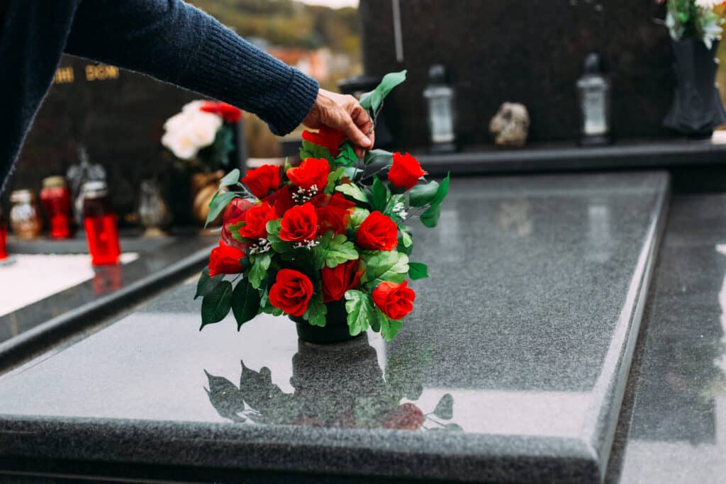 Main en pull plaçant un bouquet de roses rouges artificielles sur une tombe en granit poli. Hommage floral au cimetière.
