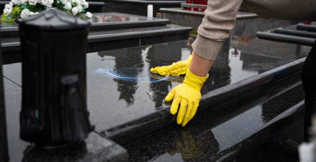Mains en gants jaunes nettoyant une pierre tombale en granit noir mouillée avec un produit bleu, des reflets visibles. Cimetière en arrière-plan.