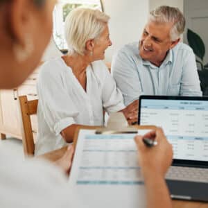 Couple senior souriant en consultation financière. Un conseiller prend des notes devant un écran affichant "HOME BUDGET".
